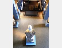 Interno di un museo visto dall’alto, con statue su piedistalli lungo i lati e una scultura centrale in primo piano su base quadrata. Sul fondo si notano una ringhiera in metallo e una scala che conduce a un livello superiore.