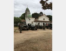 Piazzale sterrato davanti a una chiesa bianca con tetto in coppi, facciata ad archi e campanile con cupola verde e orologio. Nell’area sono parcheggiati diversi veicoli militari d’epoca, tra cui un mezzo con croce rossa, con alberi sullo sfondo sotto cielo nuvoloso.