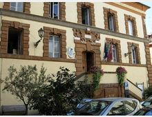 Facciata di un edificio pubblico a più piani con intonaco chiaro e cornici in pietra marrone attorno alle finestre e all’ingresso centrale sopra una scalinata. Sulla parete sono visibili uno stemma e bandiere, con fioriere ai lati e auto parcheggiate in primo piano.