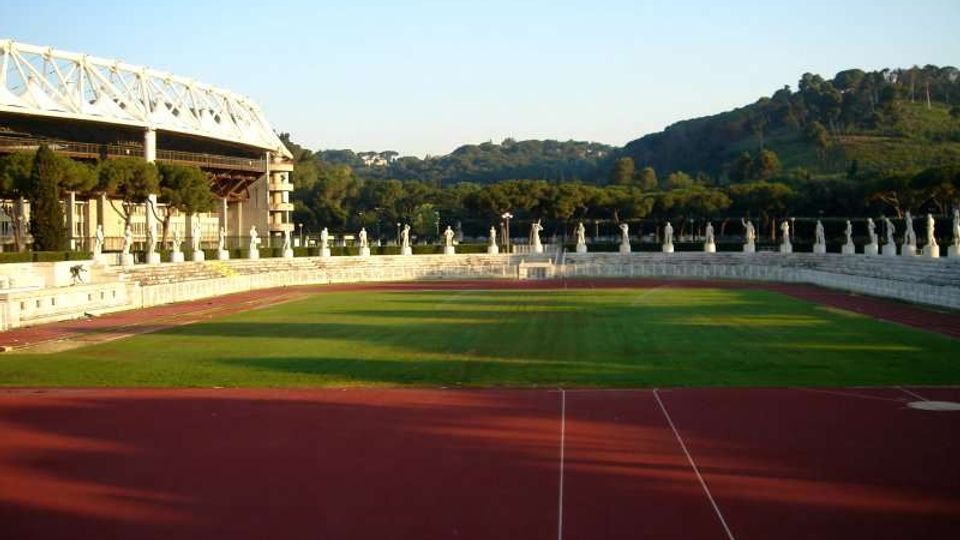 Vista di uno stadio all’aperto con pista di atletica rossa che circonda un campo in erba verde, con gradinate in cemento e una tribuna coperta sulla sinistra. Sullo sfondo si vedono colline boscose sotto un cielo sereno.