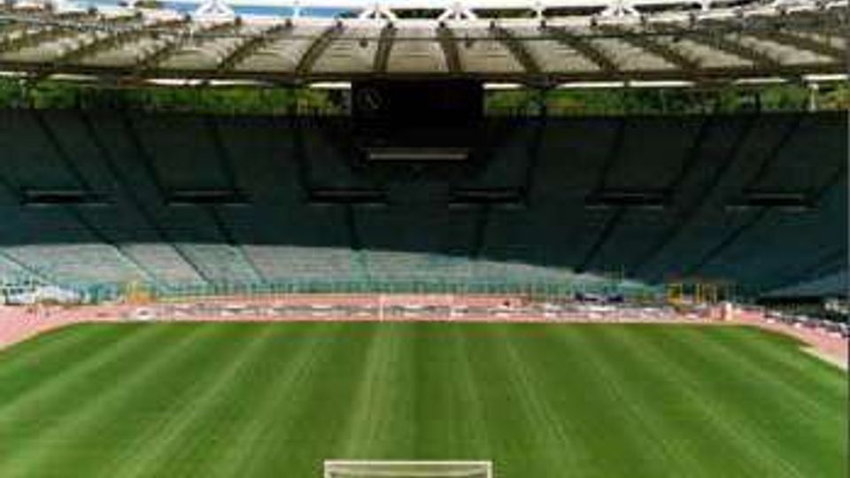 Vista dall’interno di uno stadio all’aperto con ampie tribune coperte, un grande campo in erba verde e una pista di atletica che circonda il terreno di gioco. In primo piano si vede una porta, con la copertura dello stadio e alberi sullo sfondo.