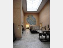 Interno di una sala museale con pareti color sabbia e lucernario a volta sul soffitto, pavimento in pietra e teche espositive con reperti lungo i lati. In fondo, una grande mappa dell’Italia dipinta sulla parete principale e un’ampia apertura che conduce a un’altra stanza.