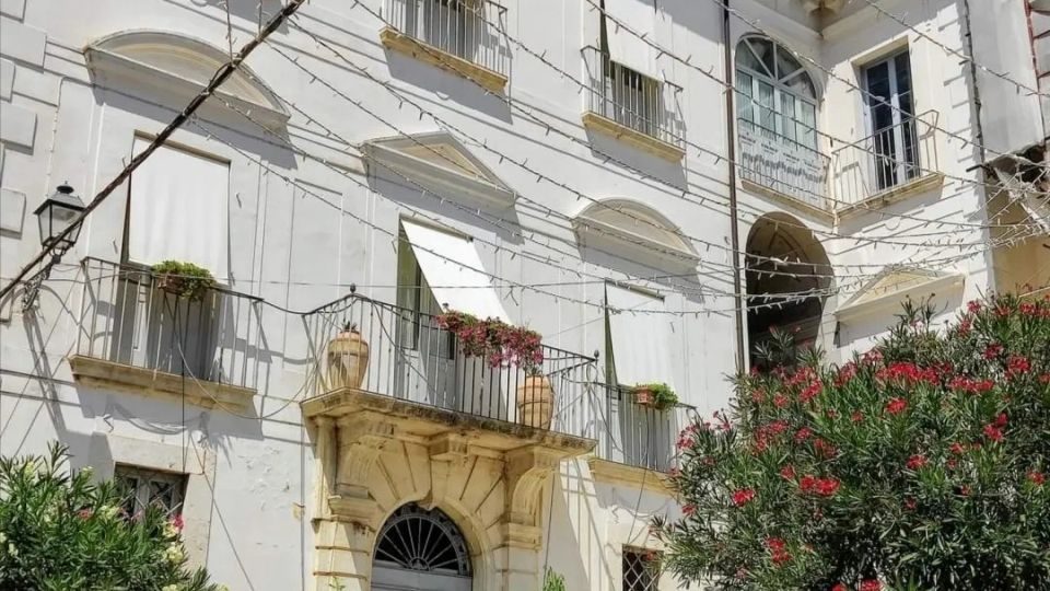 Facciata di un edificio storico intonacato di bianco, con portale ad arco in pietra, balconi con ringhiere e finestre con persiane. Nel cortile antistante si vedono fili di luminarie sospesi, vasi di fiori e alberi ornamentali con fioritura rossa.