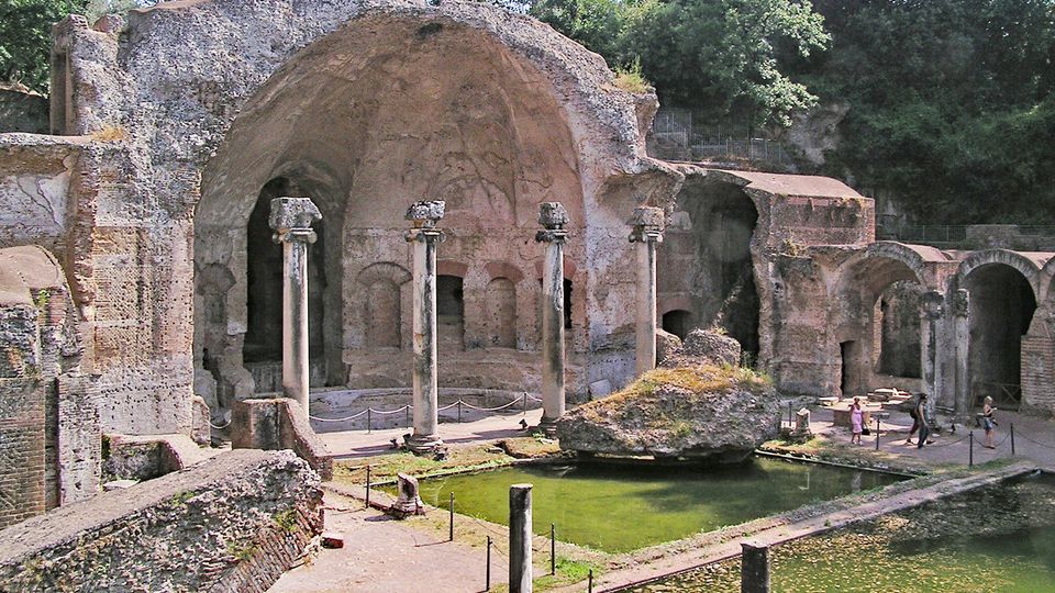 Rovine di un complesso termale romano con grandi volte in muratura, colonne in pietra e archi laterali. In primo piano si vedono vasche rettangolari con acqua verdastra e un grande masso, circondate da percorsi e aree recintate, con alberi sullo sfondo.