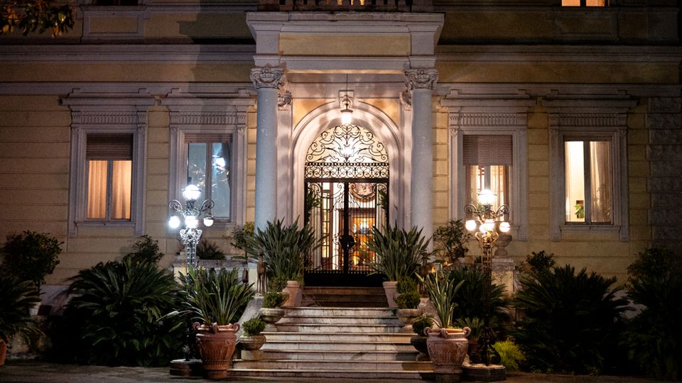 Facciata notturna di una villa storica con ingresso centrale ad arco, portone in ferro battuto e scalinate in pietra. Colonne, balcone sopra l’entrata, finestre illuminate e giardino con piante in vaso e lampioni ai lati incorniciano l’accesso.