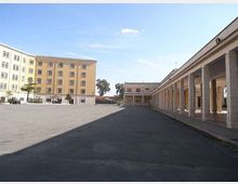 Ampia piazza asfaltata con edifici rettangolari a più piani sul lato sinistro e un lungo portico colonnato con copertura sul lato destro. Sullo sfondo si vedono altri edifici bassi e alcuni alberi sotto un cielo sereno.