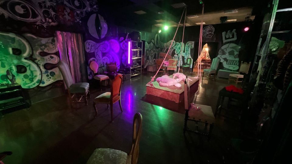 Interno di uno spazio scenico tipo teatro o studio con pavimento scuro lucido, illuminazione colorata verde e viola e pareti ricoperte da grandi murales in stile street art. Al centro c’è una pedana con corde sospese e cuscini, circondata da sedie vintage e da alcuni elementi d’arredo e luci da palco.