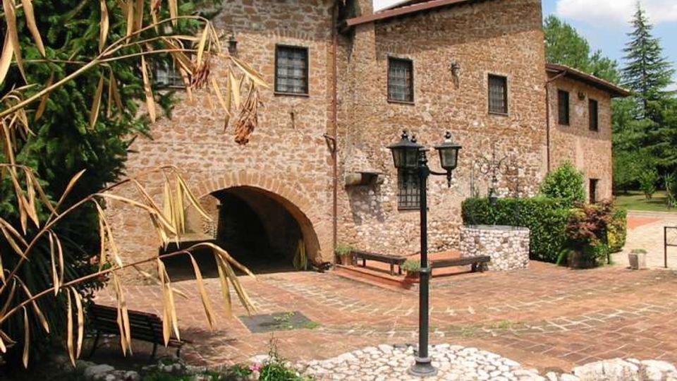 Edificio storico in pietra con finestre piccole e tetto in coppi, affacciato su un cortile lastricato con un grande arco d’ingresso. Nel cortile si vedono un lampione nero su base in ciottoli, panchine e aiuole con siepi e alberi sullo sfondo.