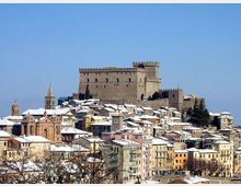 Veduta panoramica di un borgo collinare con case compatte dai tetti innevati e una grande rocca in pietra merlata in cima. In primo piano si distinguono edifici colorati e una chiesa con campanile, sullo sfondo cielo limpido.