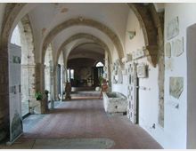 Corridoio interno in stile storico con archi in pietra e soffitto a volte, pavimento in mattoni e pareti bianche. Lungo le pareti sono esposti pannelli informativi e rilievi in pietra, con una grande vasca in pietra sul lato destro.