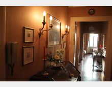 Corridoio interno con pareti color ocra illuminate da applique a candelabro, quadri incorniciati e un mobile scuro con vassoio, fiori e candelabri. In fondo si apre una stanza con pavimento in legno, finestra luminosa e un tavolino d’appoggio vicino all’ingresso.