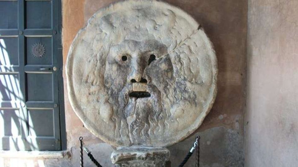 Mascherone circolare in pietra con volto scolpito e apertura della bocca, montato su un piedistallo all’interno di un ambiente in muratura intonacata. A sinistra si vede una finestra con persiane verdi e una catena di protezione delimita l’area davanti alla scultura.