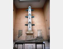 Sala museale con pareti color ocra e lucernario, dominata da una colonna marmorea alta con elementi metallici decorativi sporgenti. In primo piano è esposto un lungo reperto lapideo su supporti, con bassorilievi e targhe informative alle pareti.