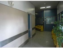 Corridoio interno di una struttura pubblica con pavimento in graniglia, pareti chiare con fascia grigia e orologio a parete. In fondo si vede una zona d’attesa con sedute gialle, pannelli e porte blu, illuminazione a neon e una pianta lungo il lato destro.