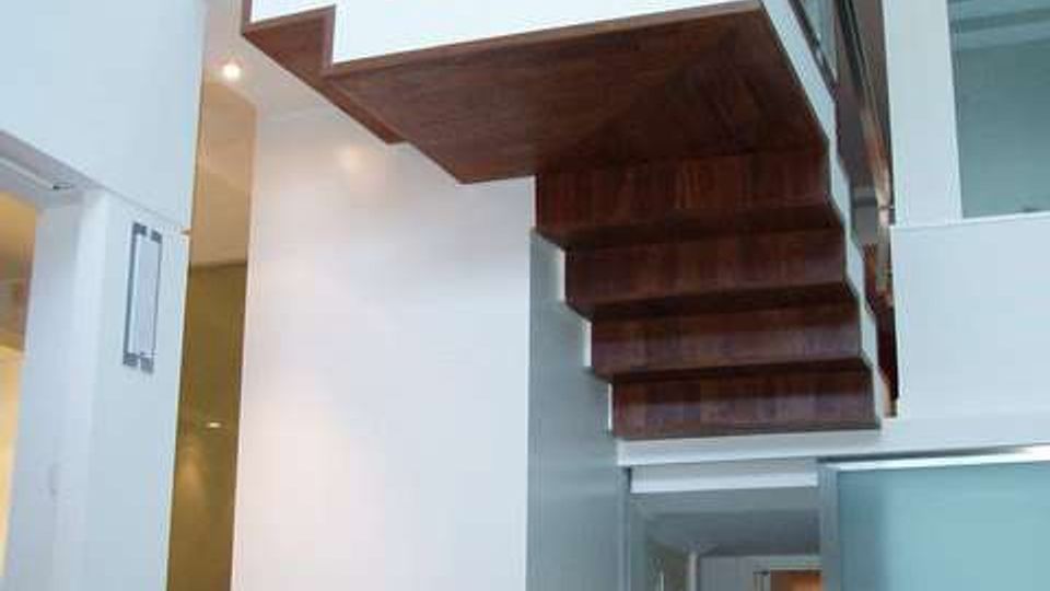 Interno moderno a doppia altezza con scala a sbalzo in legno e ringhiera in metallo che conduce a un soppalco. Pareti bianche, pavimenti in vetro e un camino incassato completano l’ambiente.