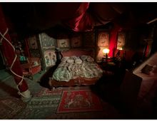 Interno di una camera da letto in stile antico con grande letto in legno e coperta di pelliccia, cuscini decorativi e tappeti ornamentali. Le pareti sono rivestite con pannelli dipinti e tende rosse drappeggiate, con lampade soffuse e mobili vintage ai lati.