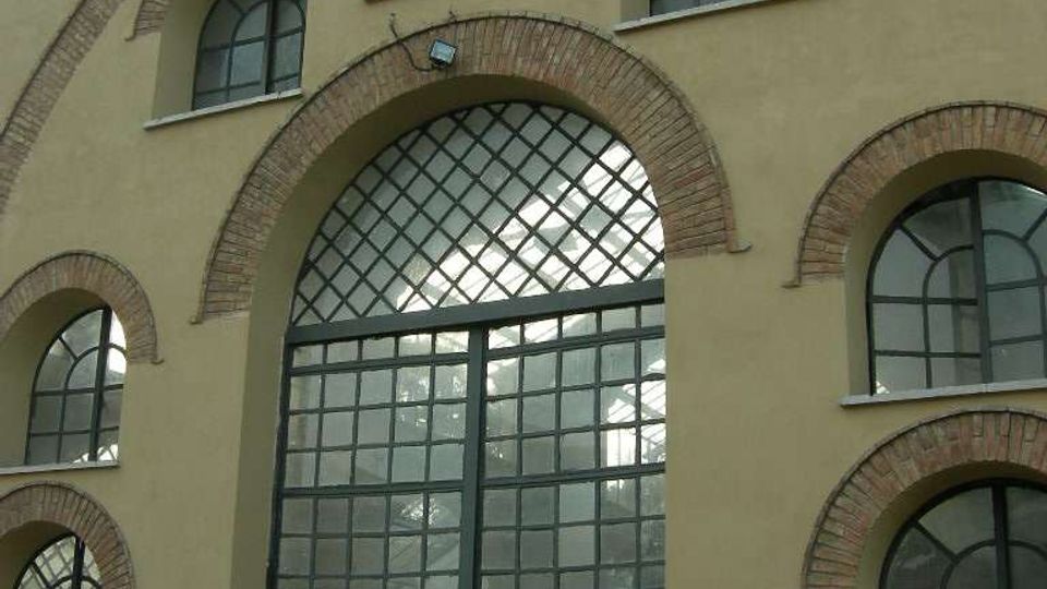 Facciata di un edificio industriale con grandi finestre ad arco incorniciate in mattoni e una porta centrale vetrata a griglia sotto un arco maggiore. Intonaco beige, struttura simmetrica con più aperture ad arco e recinzione metallica in primo piano.