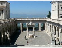 Cortile interno in pietra di un complesso monumentale con porticati ad archi sui lati e balaustre superiori. Al centro si vede un pozzo con colonne e, ai lati, statue su piedistalli; sullo sfondo panorama collinare e montagne.