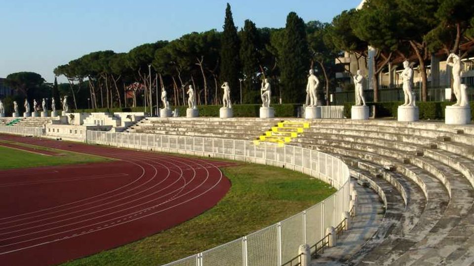 Gradinate in pietra di uno stadio all’aperto con pista di atletica rossa e recinzione metallica lungo il bordo. Sul coronamento delle gradinate si allinea una serie di statue bianche su piedistalli, con alberi alti sullo sfondo e una struttura coperta sulla destra.