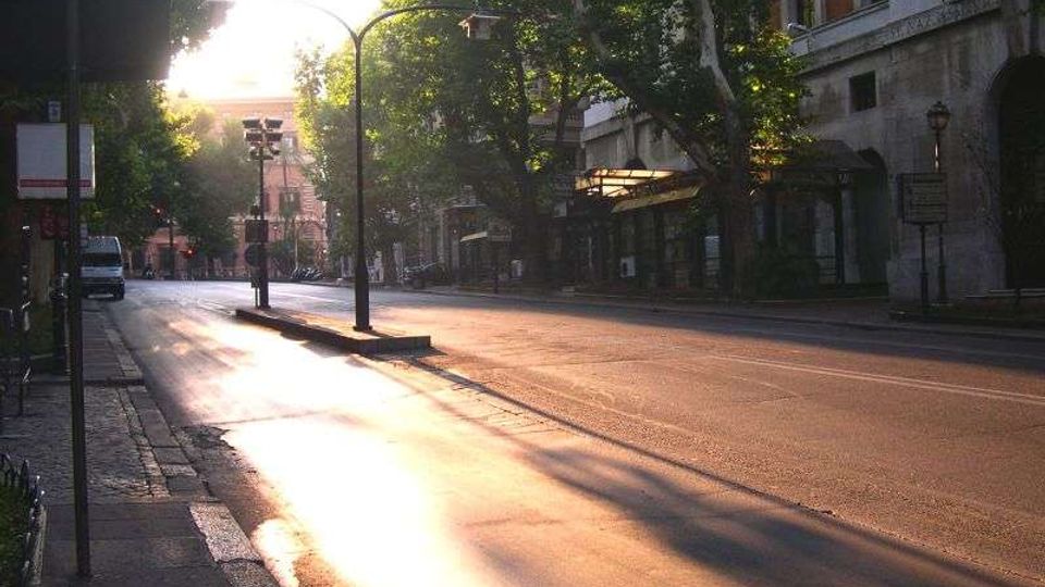 Ampio viale urbano con carreggiata asfaltata quasi vuota, fiancheggiato da grandi alberi e lampioni. Ai lati si vedono marciapiedi e facciate di edifici, con luce del sole bassa che illumina la strada in lontananza.