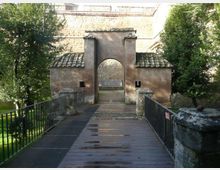 Ponte pedonale in legno con ringhiere metalliche che conduce a un ingresso ad arco in muratura, affiancato da due piccoli edifici con tetti in coppi. Sullo sfondo si vede una grande struttura in pietra, circondata da alberi e vegetazione.