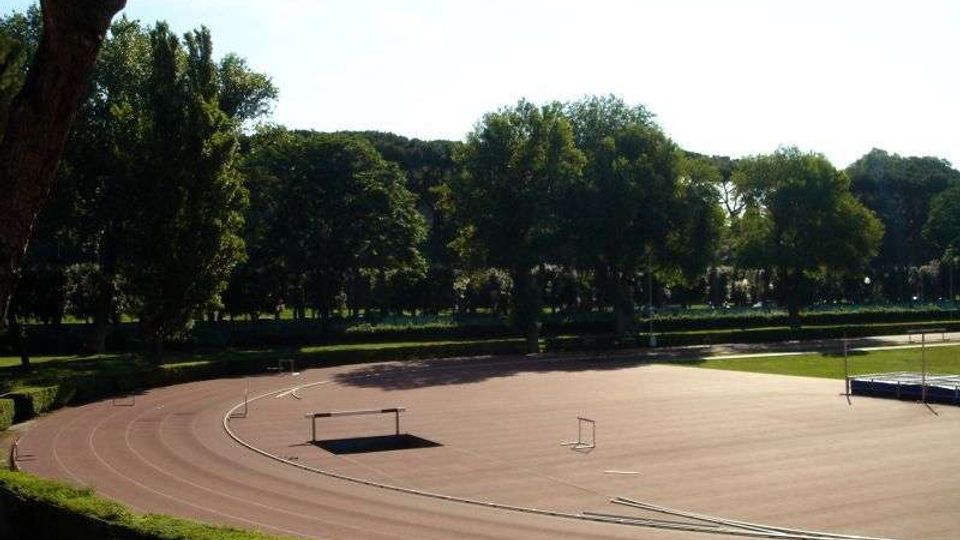 Pista di atletica all’aperto con curve marcate e alcune barriere per l’allenamento, circondata da siepi e da un’area verde. Sullo sfondo si vede una fila di alberi alti che delimita il parco sotto un cielo sereno.