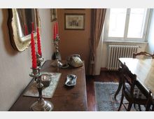 Interno di una sala da pranzo con tavolo in legno e sedie, finestra con tende e termosifone sotto. In primo piano una credenza con candelabri in argento e candele rosse, telefono d’epoca, specchio e quadri alle pareti, pavimento in legno con tappeto.