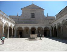 Cortile interno in pietra di un edificio storico con portici ad arcate su tre lati e facciata centrale con frontone e finestra rettangolare. Al centro si trova un pozzo o basamento in pietra su pedana rialzata, circondato da ampio pavimento lastricato.