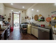 Cucina abitabile con pareti rivestite di piastrelle chiare a motivo geometrico e pavimento in cotto, con tavolo centrale e mobili lungo le pareti. Porta-finestra aperta su un piccolo balcone con sedie e vista su alberi, e zona lavello con lavastoviglie a vista.