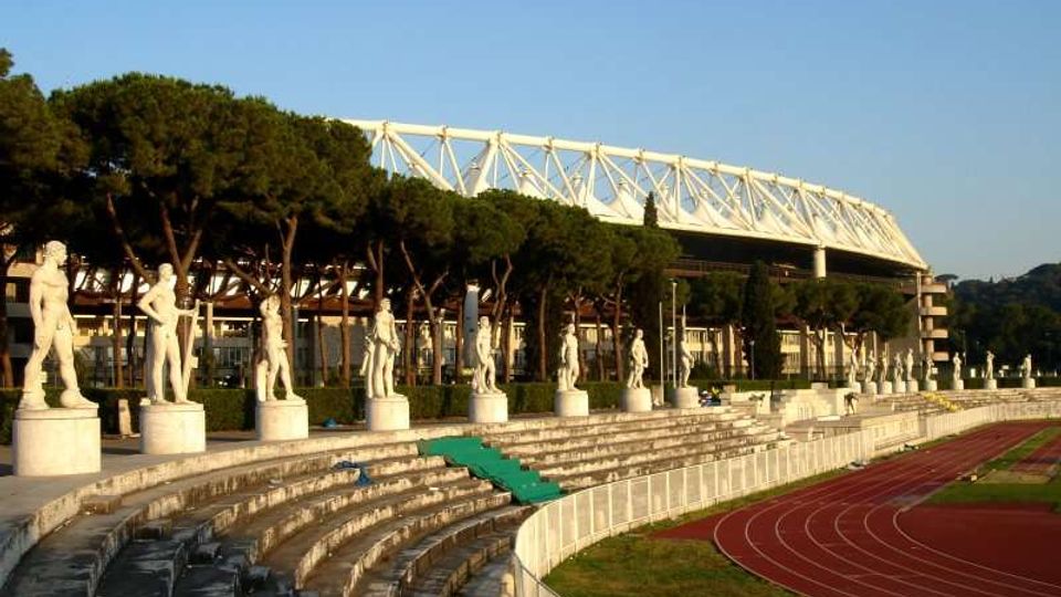 Stadio all’aperto con gradinate in pietra disposte a semicerchio, fiancheggiate da statue su piedistalli lungo il bordo superiore. In primo piano si vede una pista di atletica rossa e, sullo sfondo, una grande tribuna coperta con struttura metallica bianca e alberi alti dietro le gradinate.