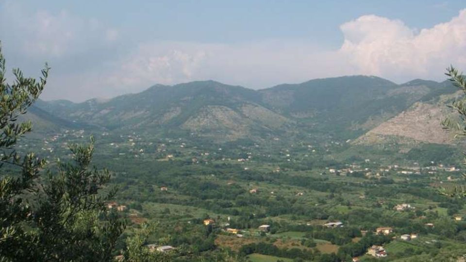 Veduta panoramica di una valle rurale con campi e vegetazione fitta, punteggiata da case sparse, con catene montuose sullo sfondo. In primo piano si vedono alberi e cespugli ai margini dell’inquadratura sotto un cielo parzialmente nuvoloso.