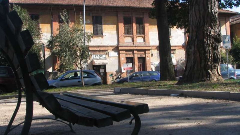 Panchina in legno e metallo in primo piano in un’area verde urbana con grandi alberi e vialetto. Sullo sfondo si vede una strada con auto parcheggiate e un edificio a due piani in laterizio e intonaco, con persiane scure e tetto in tegole.