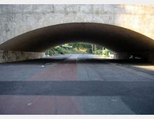 Sottopasso stradale con grande arco in pietra sopra una carreggiata ampia, con segnaletica e corsie visibili. In fondo si intravedono una strada in salita, lampioni e vegetazione ai lati.