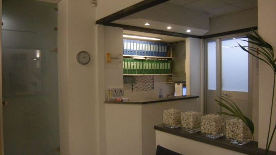 Interno di un ufficio con area reception: bancone in muratura, scaffali con raccoglitori verdi e blu e illuminazione a faretti nel soffitto. A lato si vedono una porta in vetro satinato e una finestra, con un piccolo piano d’appoggio con contenitori trasparenti.