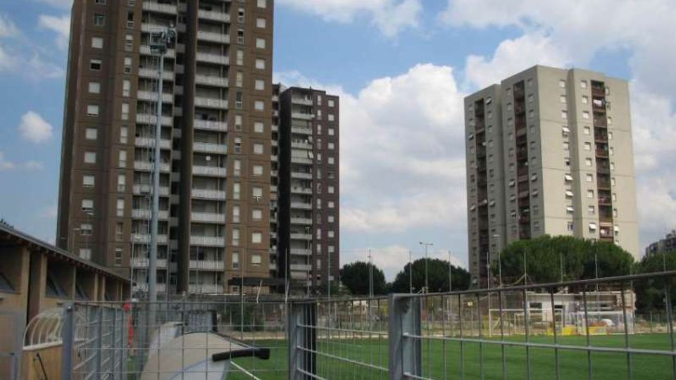 Vista urbana con due alti palazzi residenziali a più piani e balconi sullo sfondo. In primo piano si vede un’area verde recintata con reti metalliche e un passaggio pedonale, sotto un cielo parzialmente nuvoloso.
