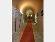 Lungo corridoio in pietra con soffitto a volta illuminato da applique, pavimento in mattoni e passatoia rossa centrale. Sulle pareti sono visibili armi e stemmi decorativi, con finestre ad arco su un lato e una porta in fondo.