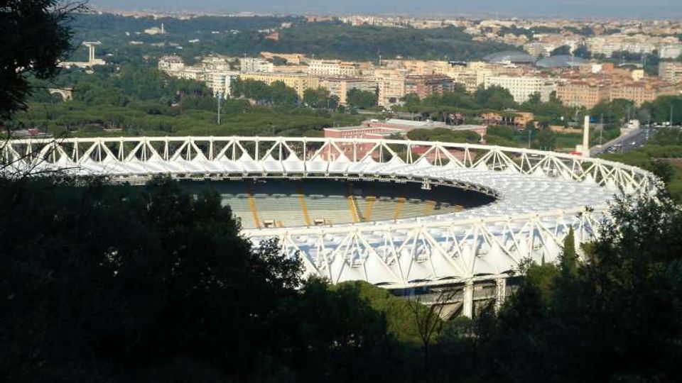 Veduta panoramica di un grande stadio ovale con struttura esterna bianca a traliccio, circondato da alberi e aree verdi. Sullo sfondo si estende la città con edifici e strade, osservata da un punto rialzato.