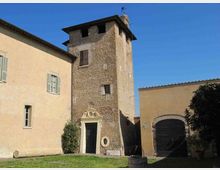 Cortile con prato davanti a un edificio storico in pietra con torre rettangolare e piccolo portale decorato. Ai lati si vedono corpi di fabbrica intonacati con finestre e un grande portone ad arco, sotto un cielo sereno.