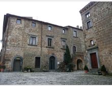 Piazza lastricata in pietra circondata da edifici storici in muratura di pietra, con finestre rettangolari e portali ad arco. Al centro si vedono una piccola scalinata, un arco passante e vasi con piante vicino agli ingressi.