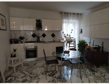 Cucina moderna con pensili bianchi lucidi e piano di lavoro lineare con fornelli e forno; parete piastrellata bianca con inserti a rombo neri. Al centro c’è un tavolo rotondo in vetro con sedie nere, pavimento effetto marmo e una grande finestra con tende leggere accanto a una credenza.