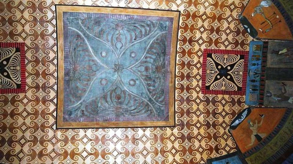 Soffitto decorato con motivi geometrici ripetuti in oro e crema e un grande riquadro centrale blu con disegno simmetrico a quattro petali. Ai lati si vedono pannelli quadrati e fasce curve dipinte con ornamenti e figure stilizzate lungo il bordo.