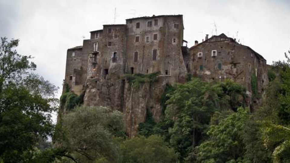 Castello medievale in pietra arroccato su uno sperone roccioso, con mura alte e piccole finestre. Intorno, vegetazione fitta di alberi e arbusti sotto un cielo nuvoloso.