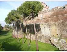 Mura di una fortezza in mattoni e pietra viste dall’esterno, con un ampio fossato erboso ai piedi. Alcuni alti pini costeggiano il muro, che presenta superfici consumate e una struttura massiccia sullo sfondo.