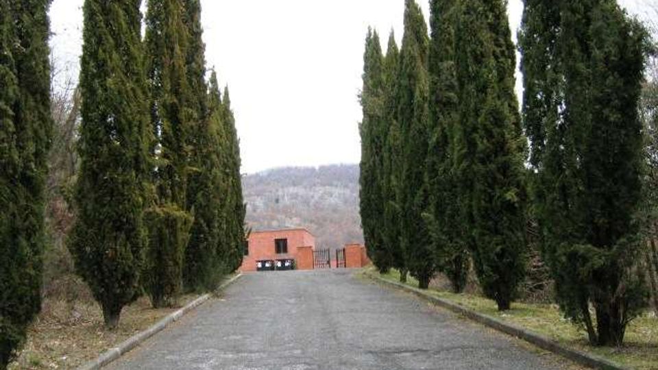 Viale asfaltato in leggera discesa fiancheggiato da alti cipressi su entrambi i lati. In fondo si vede un edificio basso in mattoni e, oltre, colline sullo sfondo sotto un cielo nuvoloso.