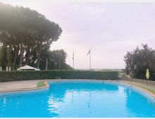Piscina all’aperto dalla forma curva con acqua azzurra e bordo in pietra, circondata da siepi e alberi alti. Sullo sfondo si vedono un gazebo, aste con bandiere e un’area paesaggistica aperta sotto un cielo nuvoloso.
