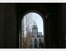 Veduta attraverso un grande arco in mattoni che incornicia un edificio con torre dell’orologio e campanile sullo sfondo. In primo piano si notano le pareti scure del portico e rami di alberi spogli contro il cielo chiaro.