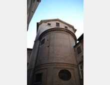 Abside semicircolare in mattoni di una chiesa storica, con cornice decorativa sotto la gronda e piccole finestre nella parte superiore. Inquadratura dal basso in una stretta via tra edifici, con cielo sereno sullo sfondo.