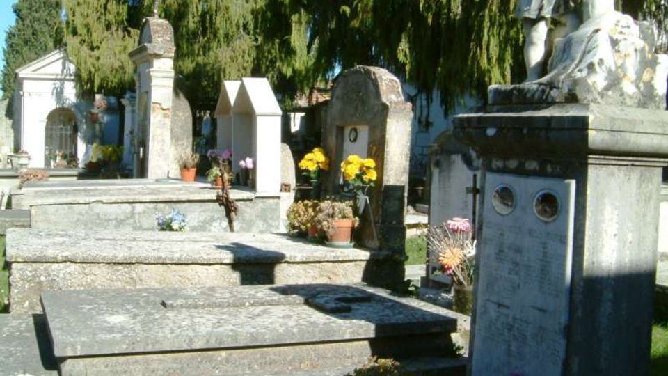 Cimitero con tombe e monumenti funerari in pietra disposti in file, alcune con croci e vasi di fiori. Sullo sfondo si vedono cappelle e alti alberi sempreverdi che ombreggiano l’area.