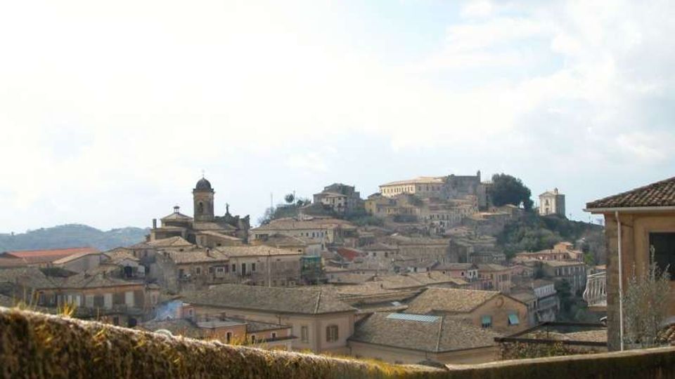 Veduta panoramica di un borgo collinare con fitte case in pietra dai tetti in coppi e una chiesa con campanile al centro. Sullo sfondo si distinguono edifici storici più grandi sulla sommità della collina, sotto un cielo parzialmente nuvoloso.