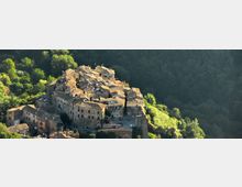 Veduta aerea di un borgo medievale arroccato su una collina, con case in pietra addossate tra loro e tetti in tegole. Il paese è circondato da un fitto bosco verde e si nota una cinta muraria lungo il margine dell’abitato.